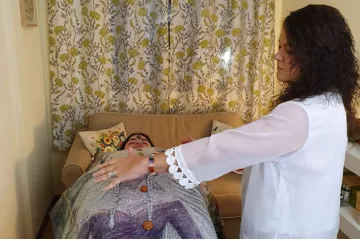 Persona recibiendo sesión de Reiki en una camilla con una manta, mientras un terapeuta de pie extiende las manos sobre ella, en un entorno acogedor con almohadas y una cortina floral de fondo.